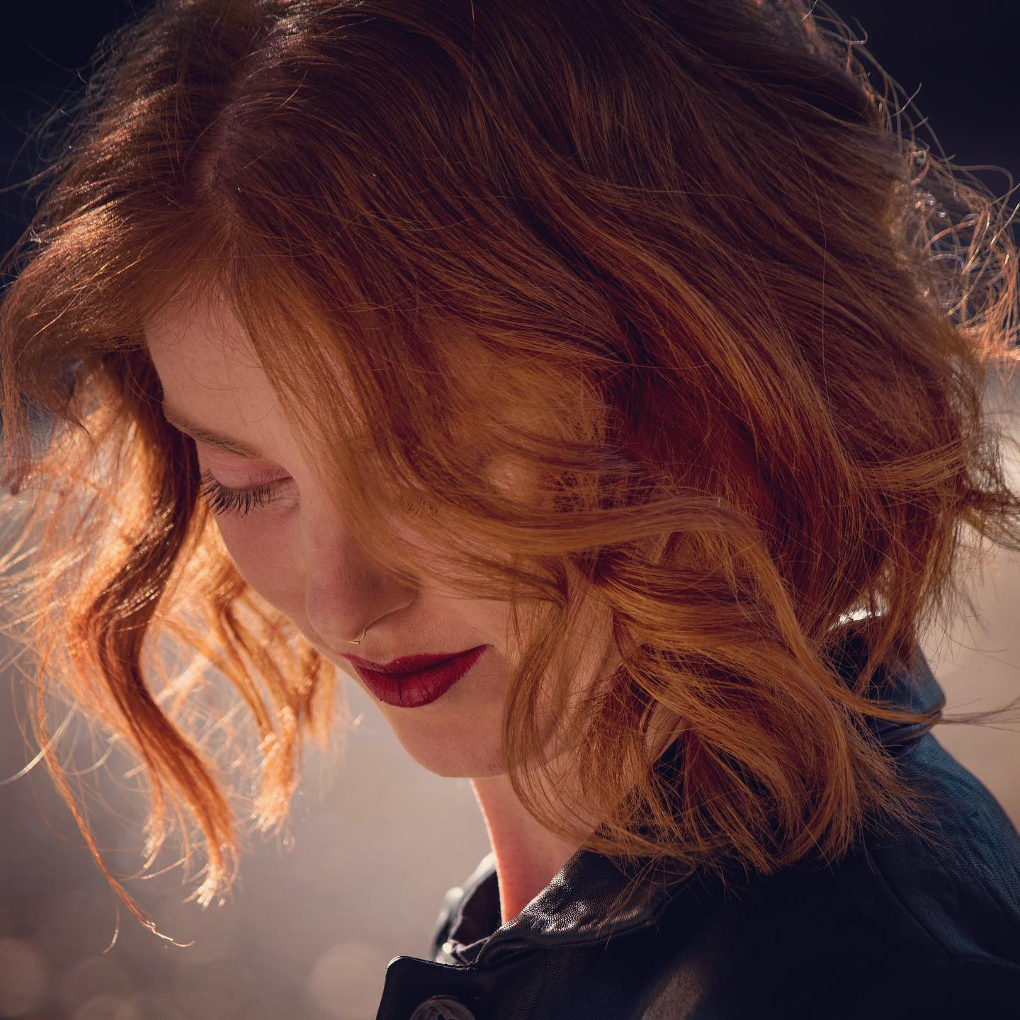 Woman with a vibrant, medium-length copper hairstyle featuring loose waves and soft layers, illuminated by backlighting, creating a warm, glowing effect
