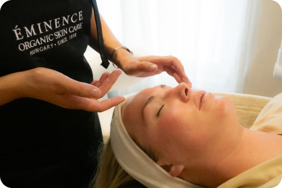 A skincare professional in an Eminence Organic Skin Care apron provides a relaxing facial treatment to a client lying down with eyes closed.
