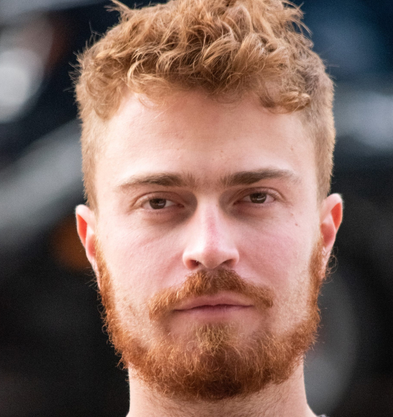 Man with a textured, tousled short curly hairstyle and a full beard, looking intently forward