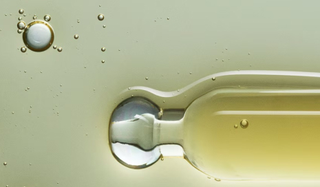Close-up of an oil dropper, with a single drop about to fall, set against a background of light yellow oil with small bubbles